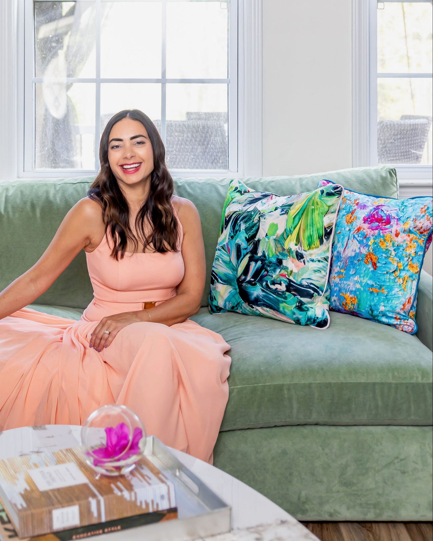 Woman in a peach dress sitting on a green couch with colorful pillows in a bright room.